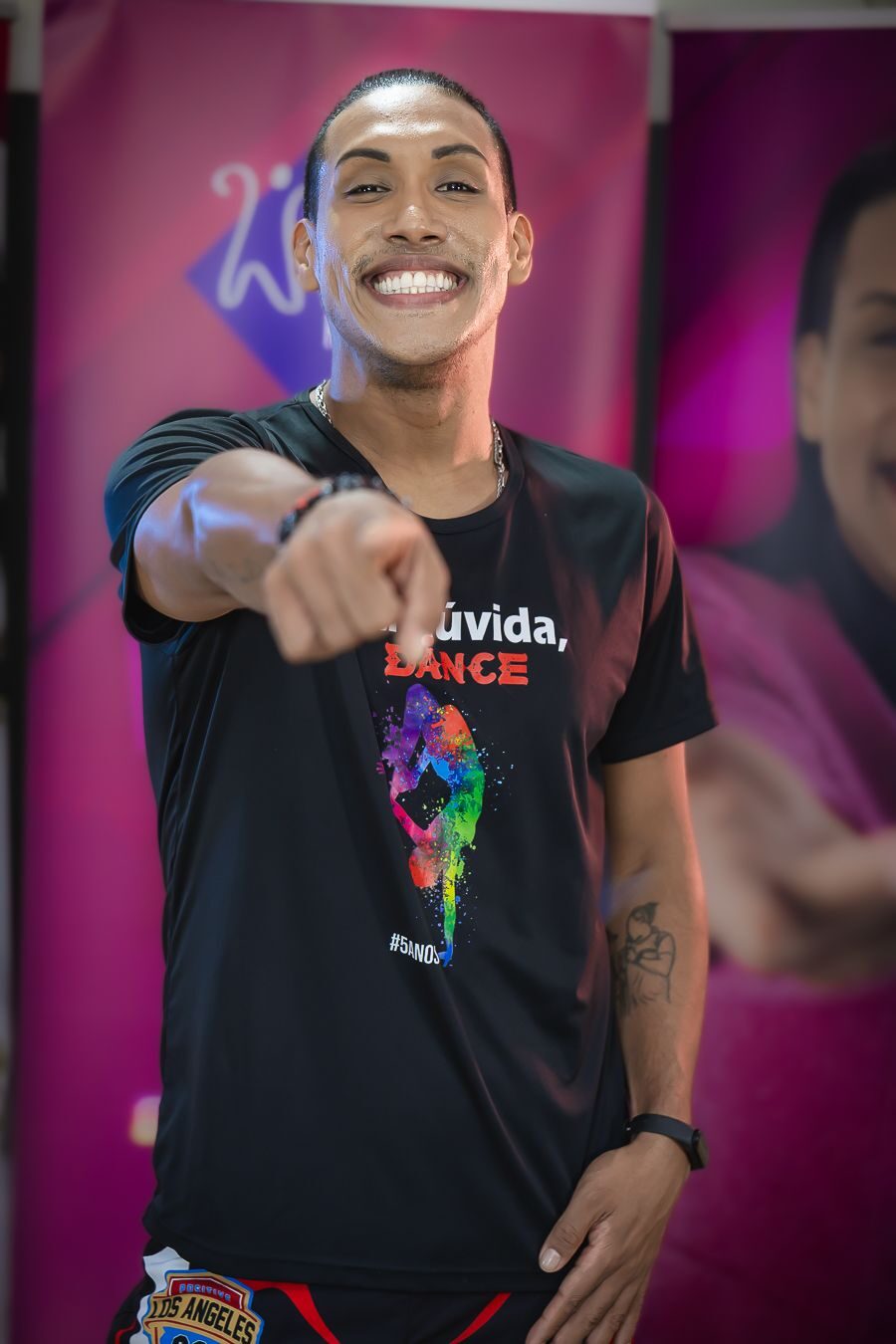 Walace Rocha sorrindo e apontando para a câmera, vestindo a camiseta Na dúvida, DANCE